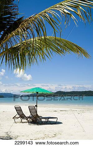 Stock Image - Deck Chairs Under Parasol On White Sandy Beach With Palm Trees Overlooking Blue Sea. Pantai Cenang (Cenang Beach), Pulau Langkawi, &#10; Malaysia, South East Asia.. Fotosearch