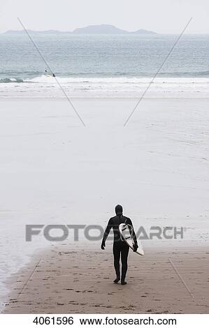 Stock Photograph - Dedicated surfer in full cover wetsuit heading out to the chilly winter waters at Whitesands Beach on the Pembrokeshire Coast Path; Pembrokeshire, Wales. Fotosearch