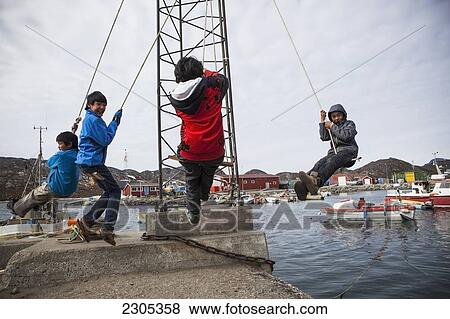 Stock Photo - Denmark, Greenland, Kids swinging over water in harbour; Paamiut. Fotosearch