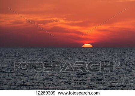 Stock Photo - Dramatic red sky with the sun setting under the ocean and horizon; Paphos, Cyprus. Fotosearch