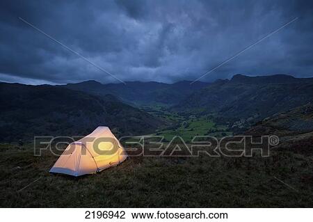Stock Image - England, Cumbria, Lit tent on hill above Langdale on stormy evening; Lake District. Fotosearch