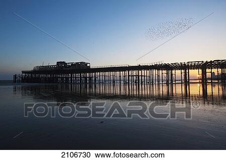 Stock Image - England, East Sussex, Ruins of Hastings pier on winter's evening with flock of starlings above; Hastings. Fotosearch
