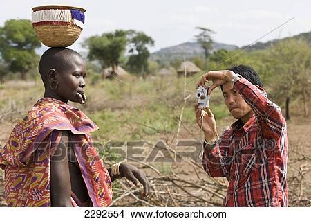 Ethiopia, Southern Nations Nationalities And Peoples' Region, South Omo, Mursiland, Japanese Tourist Taking Picture Of Mursi Tribal Woman With Pierced Lower Lip For Holding Large Pottery Lip-Plate (Dhebi); Maridungka Village View Large Photo Image Picture - Ethiopia, Southern Nations Nationalities And Peoples' Region, South Omo, Mursiland, Japanese Tourist Taking Picture Of Mursi Tribal Woman With Pierced Lower Lip For Holding Large Pottery Lip-Plate (Dhebi); Maridungka Village. Fotosearch