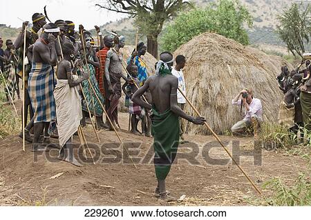 Stock Image - Ethiopia, Southern Nations Nationalities And Peoples' Region, South Omo, Southern Mursiland, American Tourist Taking Picture Of Mursi Tribal Man; Sholbi Village. Fotosearch