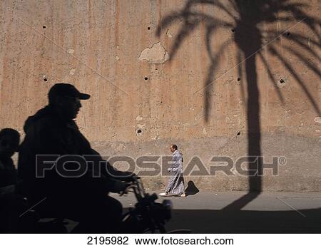 Stock Image - Father And Son On Scooter And Man In Traditional Djellaba Go Past Shadow Of Date Palm On City Walls Of Marrakesh, Morocco.. Fotosearch