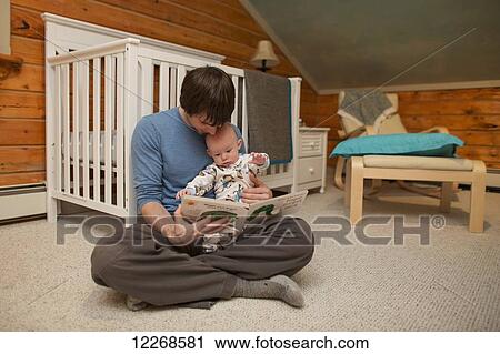 Stock Image - Father holding his child and reading a bedtime story while sitting on the floor in the nursery, USA. Fotosearch