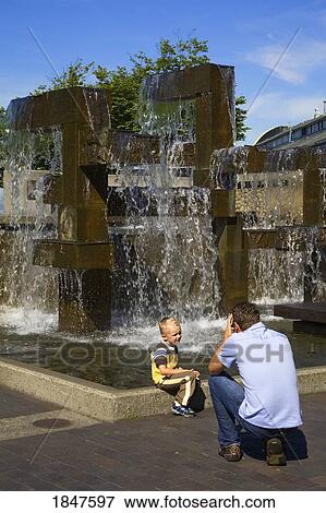 Stock Photo - Father Photographing Son At Waterfront Park Fountain; Seattle, Washington State, Usa. Fotosearch