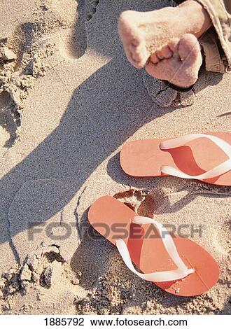 Stock Image - Feet On The Beach Next To Orange Flip Flops. Fotosearch