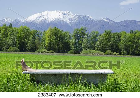 Stock Photo - Feet sticking out of a bathtub in the middle of a field with the mountains of the Swiss Alps in the background; Locarno, Ticino, Switzerland. Fotosearch