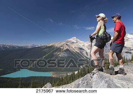 Stock Photo - Female And Male Hikers On Top Of Rock Cliff Overlooking Lake Below; Field British Columbia Canada. Fotosearch