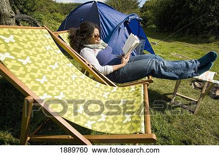 Stock Image - Female Camper Sat On Deck Chair Reading With Tent In Background. Fotosearch