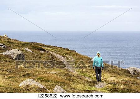 Stock Image - Female hiker along pathway of grassy hillside with rocks overlooking ocean; Ballinglanna, County Cork, Ireland. Fotosearch