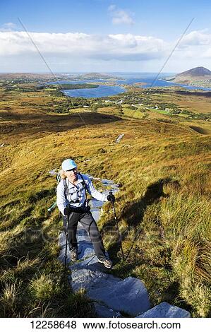 Stock Photo - Female hiker climbing trail up grassy mountain with bay in the distance; Letterfrack, County Galway, Ireland. Fotosearch