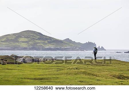 Stock Image - Female hiker on grassy field overlooking the ocean with large island in background; Omey Island, County Galway, Ireland. Fotosearch