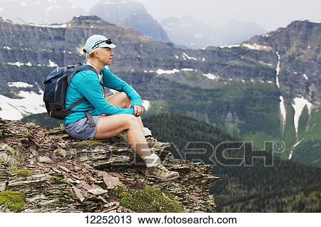 Stock Image - Female hiker sitting on a rock cliff overlooking the valley with a mountain range in the distance; Waterton, Alberta, Canada. Fotosearch