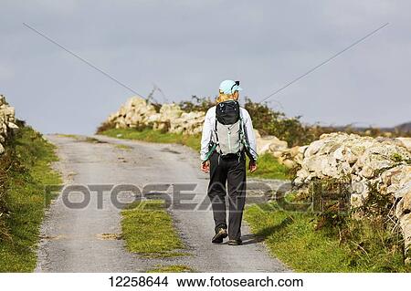 Picture - Female hiker walking along the road framed with stone fences; Omey Island, County Galway, Ireland. Fotosearch