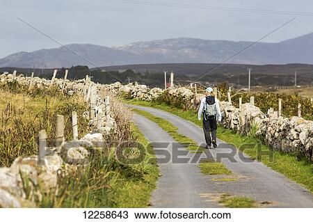 Stock Image - Female hiker walking along the road framed with stone fences and mountains in the distance; Omey Island, County Galway, Ireland. Fotosearch