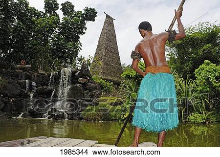 Fiji, Viti Levu, Coral Coast, Pacific Harbor, Arts Village, Fijian Man In Traditional Outfit Pushing River Raft Towards Shore With Rocks And Waterfall, Tall Bure In/Nbackground. No Model Release View Large Photo Image Picture - Fiji, Viti Levu, Coral Coast, Pacific Harbor, Arts Village, Fijian Man In Traditional Outfit Pushing River Raft Towards Shore With Rocks And Waterfall, Tall Bure In/Nbackground. No Model Release. Fotosearch