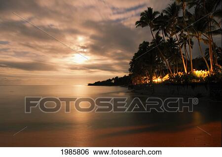 Stock Photograph - Fiji, Viti Levu, Moonlight shining brightly through clouds over calm ocean and palm lined coast; Coral Coast. Fotosearch