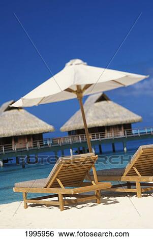 Stock Photograph - French Polynesia, Bora Bora, Beach Chairs And Umbrella With Thatched Bungalows, Blue Sky And Water. Fotosearch