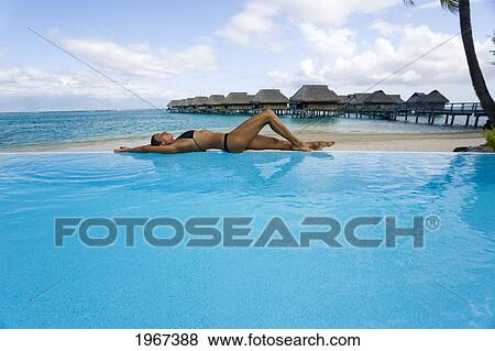 Stock Photo - French Polynesia, Moorea, Woman Relaxing At Resort Poolside, Luxury Resort Bungalows In Background.. Fotosearch