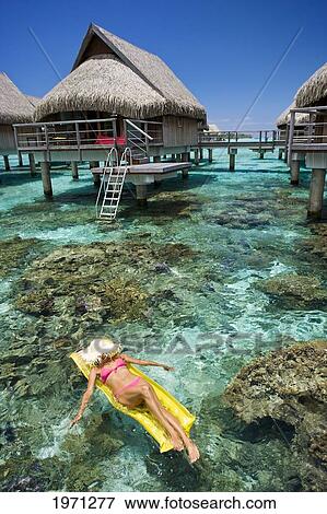 Stock Photo - French Polynesia, Moorea, Woman Sunbathing On Inflatable Raft Over Ocean Reef, Luxury Resort Bungalows Over Ocean.. Fotosearch