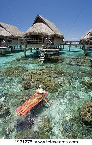 Stock Photography - French Polynesia, Moorea, Woman Sunbathing On Inflatable Raft Over Ocean Reef, Luxury Resort Bungalows Over Ocean.. Fotosearch