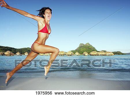 Stock Photograph - French Polynesia, Tahiti, Bora Bora, Woman Leaping On The Shore With Bungalows In The Background.. Fotosearch