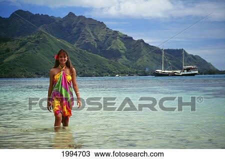 Stock Image - French Polynesia, Tahiti, Moorea, Smiling Young Woman Walks Along Shoreline In Lagoon, Flower In Hair. Fotosearch
