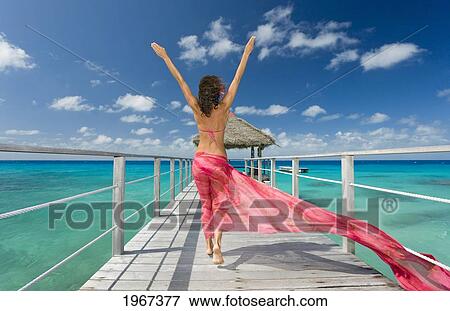 Stock Photo - French Polynesia, Tuamotu Islands, Rangiroa Atoll, Woman Walking Down Ocean Pier Wearing Pink Pareo, View From Behind.. Fotosearch