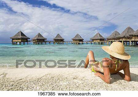Stock Photography - French Polynesia, Tuamotu Islands, Rangiroa Atoll, Woman Lounging On Beach, Luxury Resort Bungalows In Background.. Fotosearch
