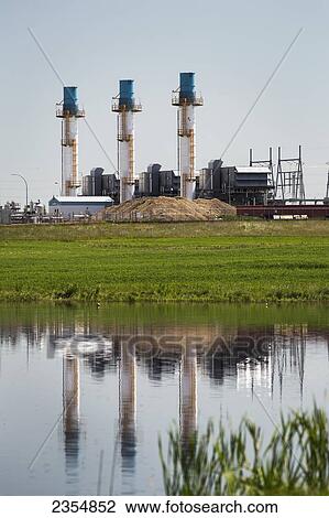 Stock Image - Gas Plant In A Green Field With Three Tall Stacks Reflecting In A Pond With Blue Sky; Crossfield, Alberta, Canada. Fotosearch