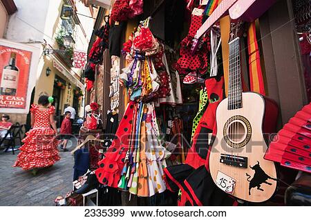 Stock Photo - Girl In Traditional Seville Dress Passing Shop Selling Souvenirs In Old Town; Santa Cruz, Seville, Andalucia, Spain. Fotosearch