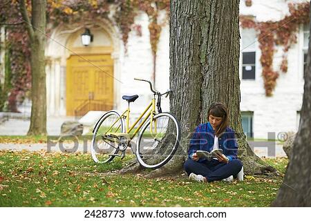 Stock Image - Girl reading book on university campus in fall with yellow bicycle; Kingston, Ontario, Canada. Fotosearch