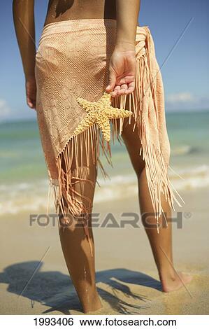 Stock Photograph - Girl Standing On Tropical Beach Wearing Pareo, Holding A Starfish, Legs And Torso. Fotosearch