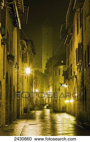 Stock Image - Golden lights illuminating buildings and a wet street; San Gimignano, Tuscany, Italy. Fotosearch