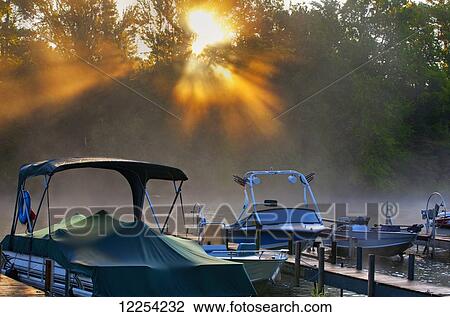 Stock Image - Golden sunlight streaming through the trees to the misty lake with boats; Foster, Quebec, Canada. Fotosearch