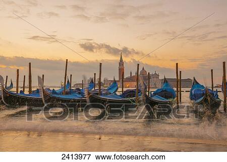 Stock Photo - Gondolas mooring at the shore of the Grand Canal with waves splashing water at sunset; Venice, Veneto, Italy. Fotosearch
