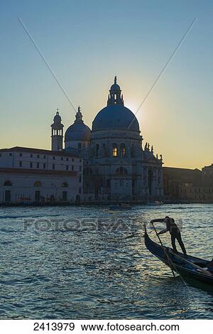 Gondolier paddles his gondola with buildings and a church along the shoreline at sunset; Venice, Veneto, Italy View Large Photo Image Stock Photo - Gondolier paddles his gondola with buildings and a church along the shoreline at sunset; Venice, Veneto, Italy. Fotosearch