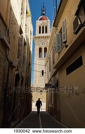 Stock Image - Greece, Crete, Man Walking Down Narrow Alley to Church; Rethymno. Fotosearch