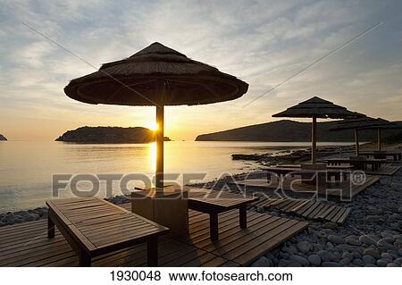 Stock Photo - Greece, Crete, Sunshade and Sun Lounger on Beach Opposite Spinalonga Island at Dawn; Spinalonga Island. Fotosearch