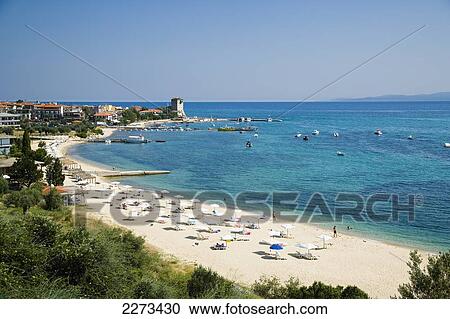 Stock Image - Greece, Halkidiki, Tourist beach with beach umbrellas and sun bathers with the Byzantine Tower or Tower of Prosforios in the distance; Ouranoupoli. Fotosearch