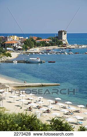 Stock Photo - Greece, Halkidiki, Tourist beach with beach umbrellas and sun bathers with the Byzantine Tower or Tower of Prosforios in the distance; Ouranoupoli. Fotosearch