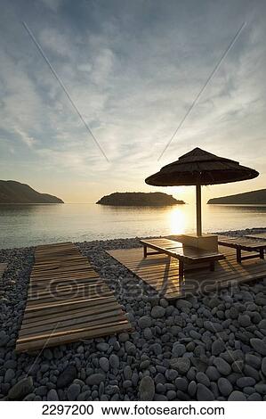 Stock Image - Greece, Umbrella and sun loungers on beach opposite Spinalonga Island at dawn; Crete. Fotosearch