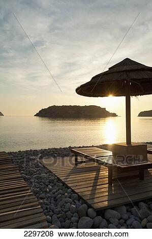 Stock Photo - Greece, Umbrella and sun loungers on beach opposite Spinalonga Island at dawn; Crete. Fotosearch