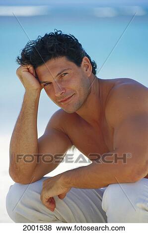 Stock Photo - Handsome Man Outdoors Wearing Long Pants, Elbow On Knee Head In Hand Shirtless Ocean Background Blur Soft Focus D1185. Fotosearch