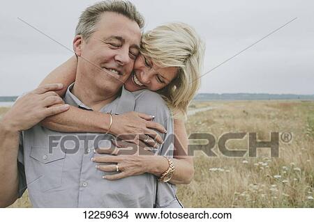 Picture - Happy senior couple embracing on a grassy beach; Coquitlam, British Columbia, Canada. Fotosearch