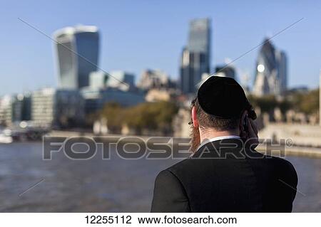Stock Image - Hasidic Jewish man on Tower Bridge overlooking the River Thames, the buildings of the City of London and the Tower of London; London, England. Fotosearch