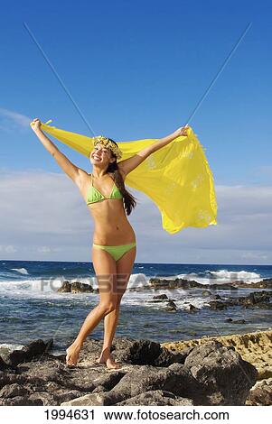 Stock Image - Hawaii, Beautiful Local Girl On Rocky Shoreline, Holding Yellow Pareo, Wearing Haku Lei. Fotosearch