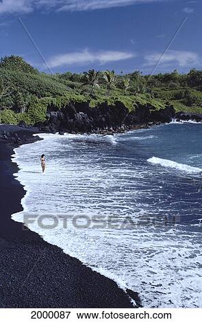 Stock Photo - Hawaii, Big Island, Woman Walks Along Shoreline Black Sand Beach, Tropical Greenery Background. Fotosearch